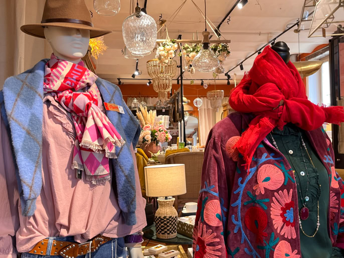 Deux mannequins vêtus de vêtements colorés, l'un avec un chapeau et une écharpe rose, l'autre avec un chapeau rouge, dans un magasin décoré avec des lampes et des objets divers.