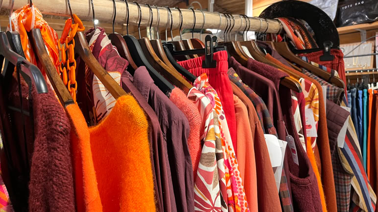 Boutique de vêtements avec des manteaux, vestes et chemises suspendus sur des portants, et des sacs à main exposés.