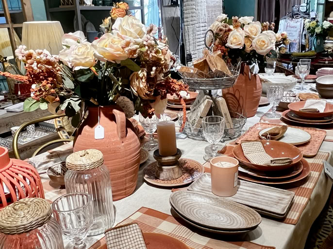Table dressée avec vaisselle en céramique, verres, bougies, et arrangements floraux dans des tons de terre cuite et beige.