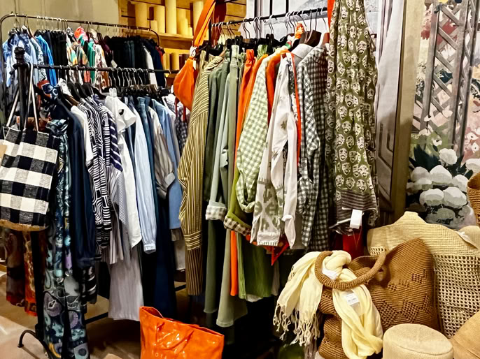 Portant de vêtements colorés avec des chemises, vestes et robes, sacs à main suspendus à gauche, et divers accessoires sur le sol.