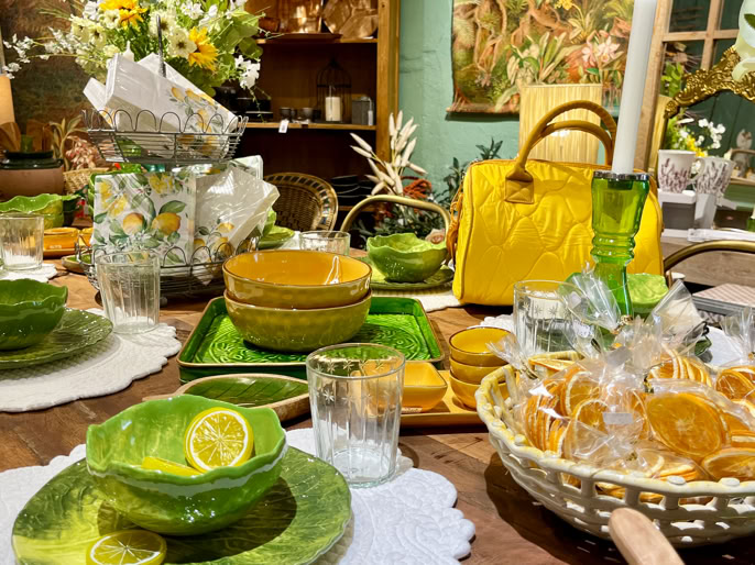 Table dressée avec vaisselle verte et jaune, bols, assiettes, verres, panier avec biscuits, sac à main jaune, bougie, et décorations florales.