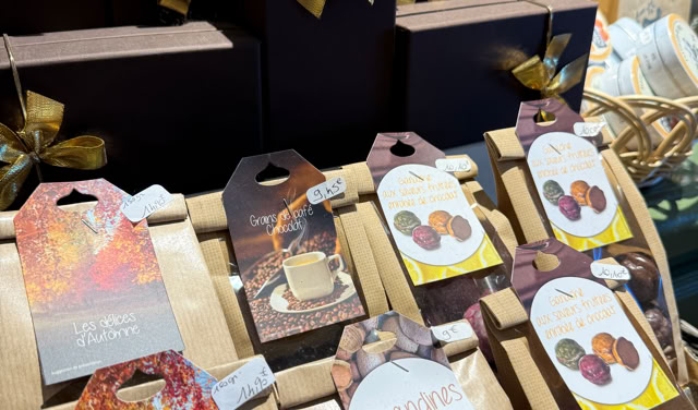 Présentoir de chocolats avec boîtes noires ornées de rubans dorés et sachets de chocolats variés, étiquettes colorées, et une personne en arrière-plan.