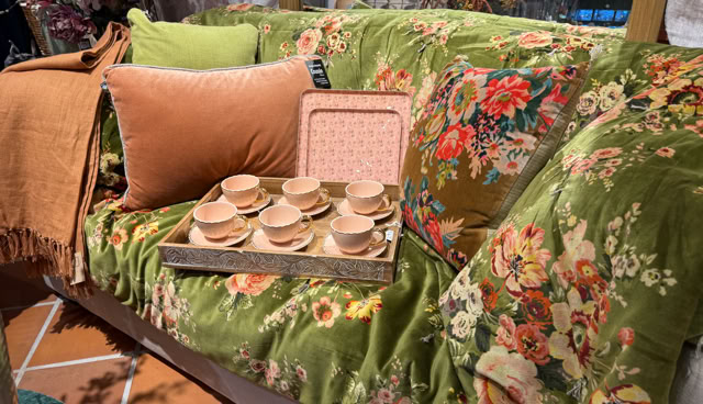 Canapé recouvert d'un tissu à motifs floraux verts, avec des coussins orange et verts, et un plateau contenant des tasses en bois.