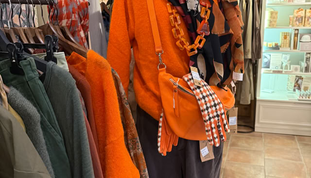 Mannequin habillé d'un pull orange, écharpe colorée, bonnet gris, et sac orange, entouré de vêtements suspendus dans un magasin.