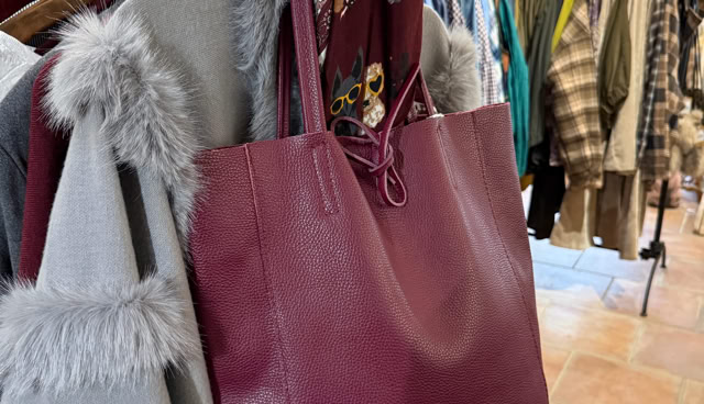 Sac à main bordeaux accroché à un portant de vêtements, avec un manteau gris à fourrure et un bonnet gris à côté.