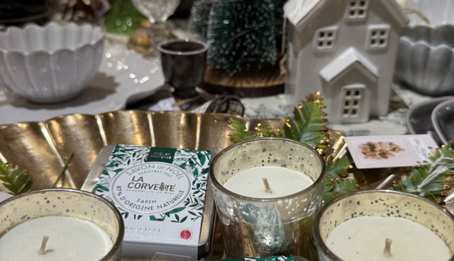 Bougies dans des verres dorés, petites boîtes avec étiquettes, miniature de sapin de Noël, et maison décorative blanche sur une table.