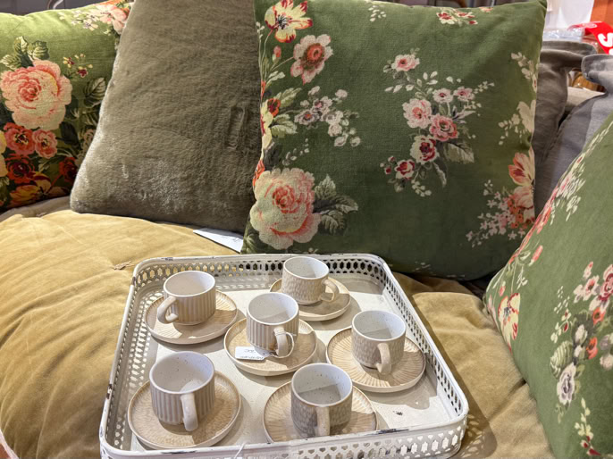 Un canapé avec des coussins verts à motifs floraux, et un plateau contenant six tasses à café blanches avec des soucoupes.