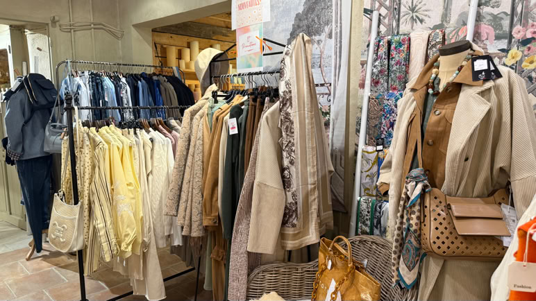 Boutique de vêtements avec des manteaux, vestes et chemises suspendus sur des portants, et des sacs à main exposés.