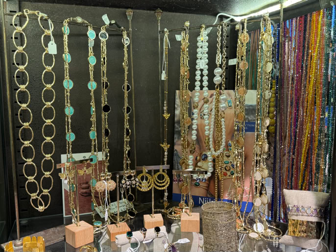 Vitrine avec divers colliers suspendus, comprenant des chaînes dorées et des perles colorées, ainsi que des flacons de parfum et des boîtes sur l'étagère inférieure.